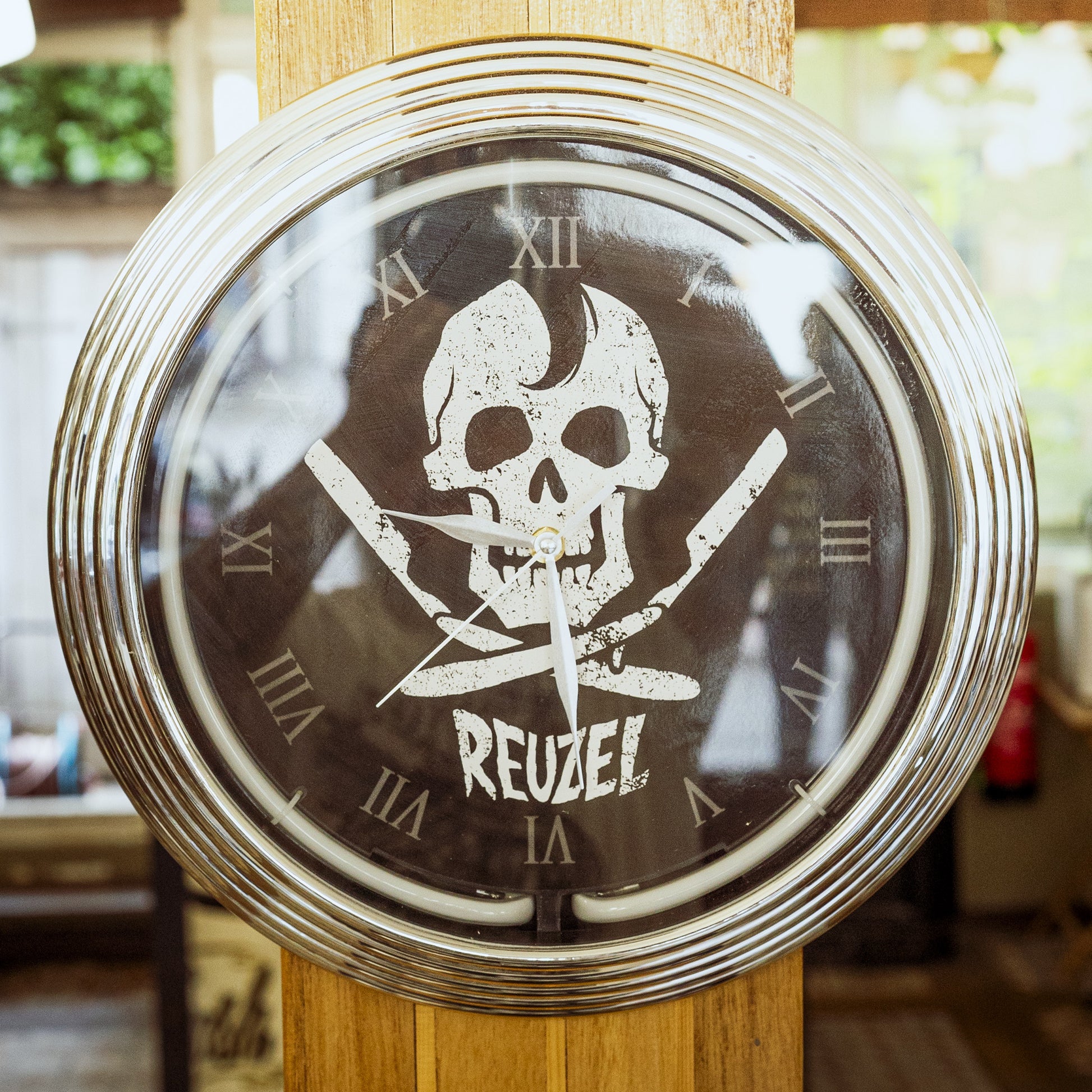 A round wall clock with a chrome silver frame and a black face featuring a distressed white skull and crossed swords graphic, with the word 'REUZEL' printed below. The clock is mounted on a wooden post in a real-world setting with a blurred background.