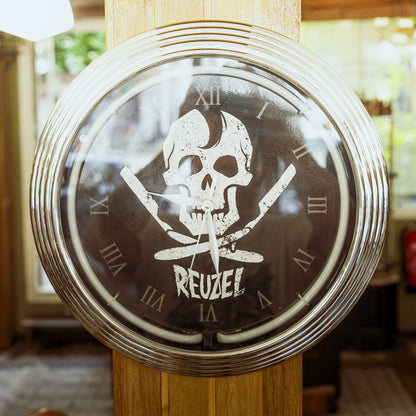 A round wall clock with a metallic silver frame and a dark face featuring a white skull and crossed knives above the word 'REUZEL'. The clock is mounted on a wooden post in a real indoor setting with blurred background details.