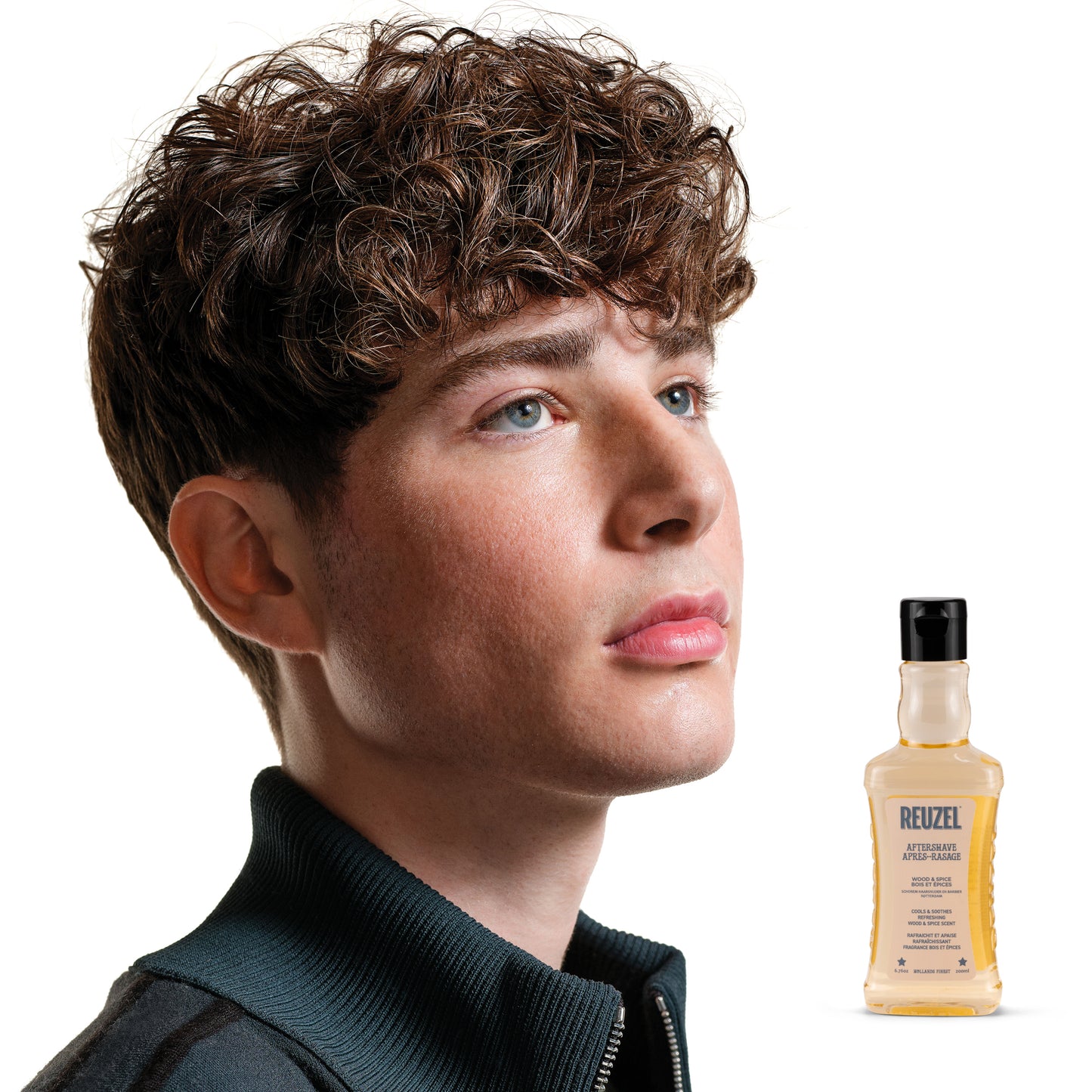 Reuzel Wood & Spice beard aftershave bottle with a black cap and label, showcasing the grooming product in a natural setting.